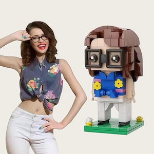 Può includere: Una figura in stile Lego con capelli castani, occhiali quadrati e un top blu con accenti floreali gialli. La figura è su una base verde. Una persona con occhiali e un top floreale è anche nell'immagine.