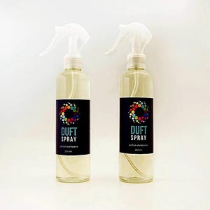 Peut inclure: Deux flacons pulvérisateurs transparents remplis d'un liquide jaune clair. Chaque flacon porte une étiquette noire avec le texte "DUFT SPRAY" et un motif circulaire coloré. Les flacons contiennent 250 ml chacun.