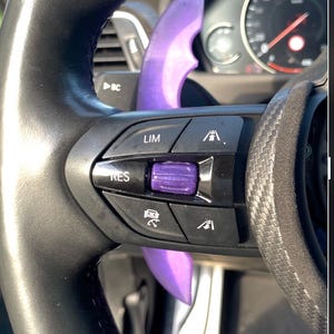 Könnte beinhalten: Nahaufnahme eines schwarzen Lenkrads mit einem violetten Akzent. Das Lenkrad hat Tasten mit der Aufschrift "LIM" und "RES". Im Hintergrund ist ein Tachometer zu sehen.