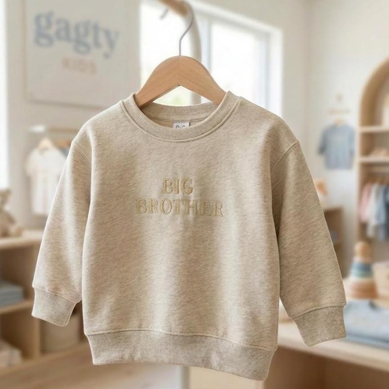 Może przedstawiać: Beżowa bluza z haftowanym napisem "BIG BROTHER". Top z długim rękawem wisi na drewnianym wieszaku. W tle znajduje się znak z napisem "gagty KIDS".