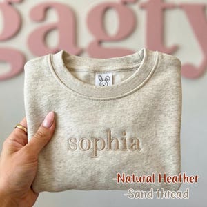 May include: A natural heather sweatshirt with the name "Sophia" embroidered in sand-colored thread. The sweatshirt features a small bunny logo and the text "Natural Heather - Sand thread".