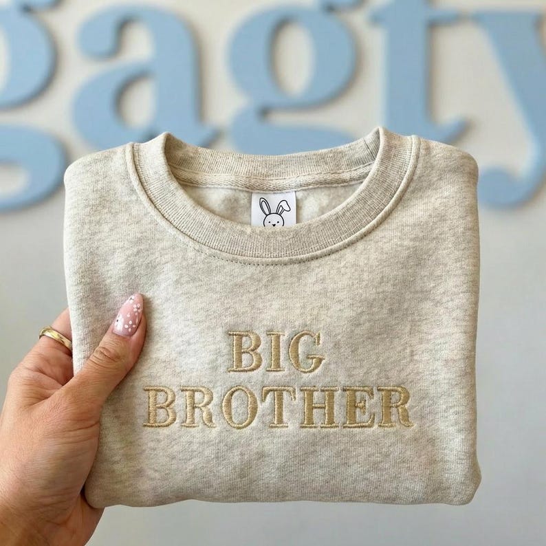 Może przedstawiać: Beżowa bluza z haftowanym napisem "BIG BROTHER" złotą nicią. Mała grafika z kr&oacute;likiem jest wyhaftowana w pobliżu kołnierza. Bluza jest złożona, z jasnoniebieskim tłem.