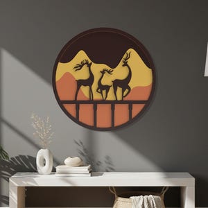 May include: A circular wall art piece featuring a layered silhouette design of three deer standing on a bridge. The background consists of stacked mountain shapes in shades of dark brown, yellow, and orange, enclosed within a dark circular frame.