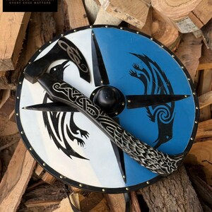 May include: A round shield with a black handle and a black axe head. The shield is divided into white and blue sections, each featuring a black raven design. The shield is trimmed with brass rivets and a black border. The handle has a carved design.