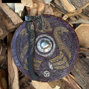 Peut inclure: Un ensemble bouclier et hache de style viking. Le bouclier est rond, avec un fond violet foncé et des motifs en relief dorés et argentés. La hache a un manche noir et une lame argentée. L'ensemble est présenté sur un fond de bois de chauffage empilé.