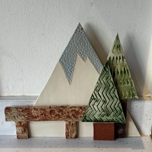 May include: A ceramic wall art piece featuring a white mountain with a textured grey peak, two green triangular trees with embossed patterns on brown trunks, and a horizontal brown textured fence-like element across the foreground.