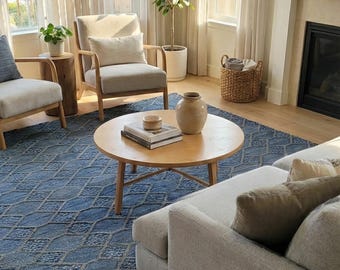 Könnte beinhalten: Wohnzimmer mit blau gemustertem Teppich, hellgrauem Sofa und zwei Sesseln. Ein runder Holztisch beherbergt Bücher und eine Vase. Transparente Vorhänge filtern das Sonnenlicht und schaffen eine helle und luftige Atmosphäre.