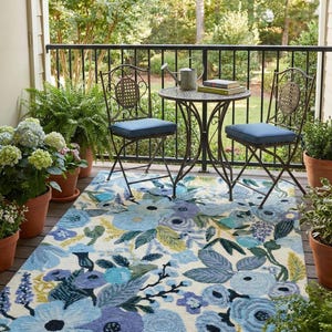 Puede incluir: Escena de patio al aire libre con una mesa y dos sillas de hierro forjado con cojines azules. Una alfombra floral con flores azules y moradas está sobre la cubierta de madera. Plantas en macetas y una barandilla negra completan la escena.