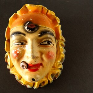 Puede incluir: Una máscara de cerámica vintage con un rostro pintado de amarillo y naranja. La máscara tiene un bigote pintado y un pequeño círculo pintado en la frente. La máscara tiene una sonrisa pintada y un pequeño círculo pintado en la nariz.