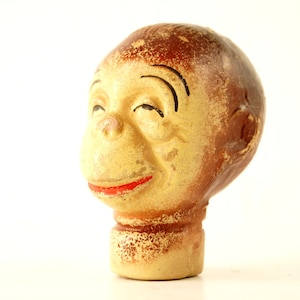Puede incluir: Una figurita de cabeza de mono de cerámica vintage con la cara pintada y el pelo marrón. El mono sonríe con los ojos cerrados y tiene la boca pintada de rojo.