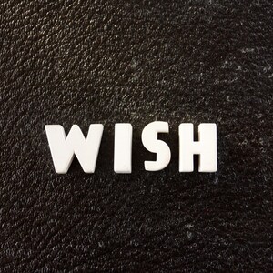 Puede incluir: La palabra "WISH" escrita en letras de plástico blancas sobre un fondo negro texturizado.