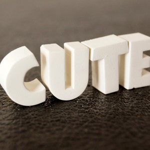 Puede incluir: Letras de madera blancas deletreando la palabra "CUTE" sobre una superficie texturizada marrón oscura.