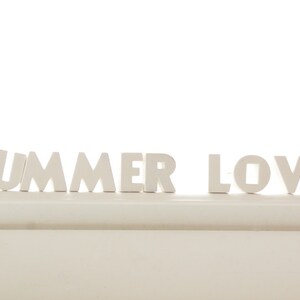 Puede incluir: Letras de madera blancas que deletrean "SUMMER LOVE" sobre una superficie blanca.