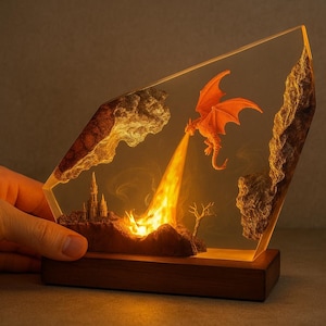 Può includere: Scultura in resina a tema fantasy con un drago arancione che sputa fuoco verso un castello. La scultura è illuminata con luce calda, posta su una base di legno.