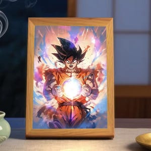 Puede incluir: Una impresión enmarcada que presenta una vibrante ilustración de un personaje de Dragon Ball Z. El personaje, con un gi naranja, está rodeado de efectos de energía, sosteniendo una esfera brillante. El marco de madera añade un toque clásico a la obra de arte.