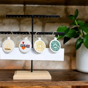 May include: Four round keychains with various designs and text, including "Garage" and "Du Bon". The keychains are white with gold key rings, displayed on a black metal stand with a wooden base. A white sign is below the keychains.