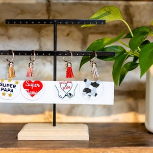 May include: A black metal earring display stand with four keychains and four round, white, graphic designs. The keychains have tassels in various colors. The designs say "Super Papa", "Super Mama", and feature hand-holding and a black and white pattern.