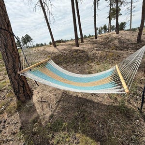 May include: A white, blue, and tan striped hammock strung between two trees. The hammock has wooden bars on each end and is suspended in a natural outdoor setting with trees and a cloudy sky.