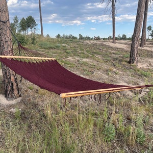 May include: A maroon hammock hangs between two trees in a wooded area. The hammock is made of a thick, woven material and has wooden spreader bars.