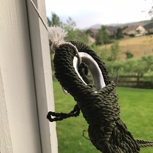 May include: Close-up of a green rope tied around a silver metal ring. The rope is tied in a knot and is secured to a white surface.
