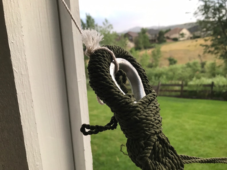 May include: Close-up of a green rope tied around a silver metal ring. The rope is tied in a knot and is secured to a white surface.