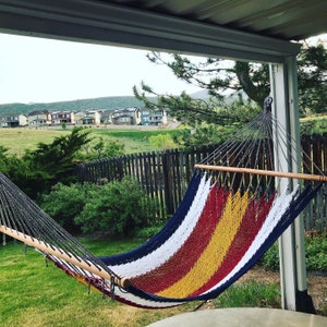 May include: A red, white, and yellow striped hammock hangs from a wooden frame under a covered patio. The hammock is made of woven cotton and has a wooden spreader bar. The hammock is in front of a wooden fence and a green lawn.