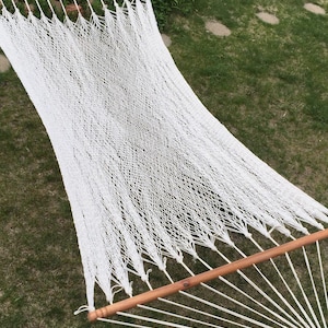 Puede incluir: Una hamaca blanca tejida con barras separadoras de madera, apoyada sobre hierba verde. La hamaca está hecha de cuerda gruesa y anudada, creando una superficie cómoda para descansar. Las barras de madera son de color marrón claro.