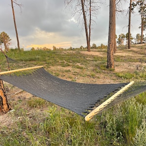 May include: A black hammock strung between two trees in a wooded area. The hammock is made of a woven material and has wooden spreader bars.