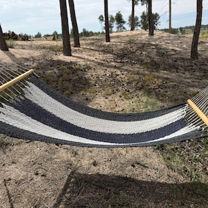 May include: A striped hammock with black, white, and gray bands, suspended between trees. The hammock has wooden spreader bars and is made of woven rope. The scene is outdoors, with a sandy ground and trees in the background.
