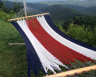 May include: A red, white, and blue striped hammock hangs between two wooden poles. The hammock is made of a woven rope material. The hammock is set up in a grassy area with a view of a mountain range in the background.