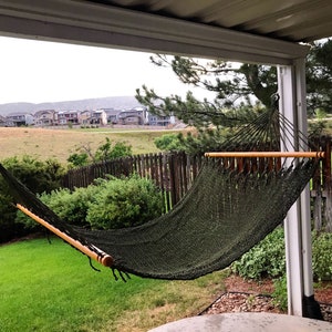 May include: A green hammock hangs between two wooden posts on a covered patio. The hammock is made of a woven material and has a wooden spreader bar. The hammock is in a backyard with a green lawn and a wooden fence in the background.
