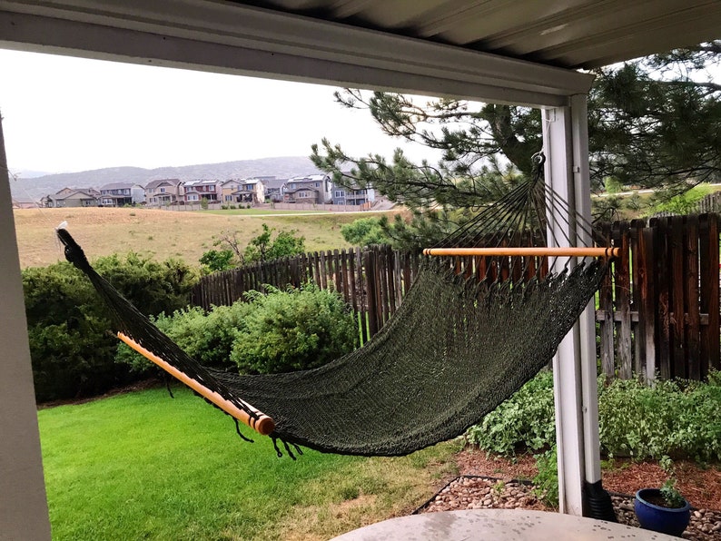 May include: A green hammock hangs between two wooden posts on a covered patio. The hammock is made of a woven material and has a wooden spreader bar. The hammock is in a backyard with a green lawn and a wooden fence in the background.