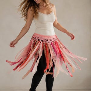 May include: A pink crochet waist wrap featuring a wide waistband with an openwork pattern and numerous hanging fabric strips in various shades of pink, red, and peach, creating a layered, fringed effect that flares outward during movement.