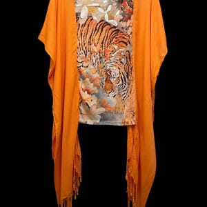 May include: A bright orange open-front shawl with fringe trim layered over a short-sleeve top. The top features a graphic print of a tiger surrounded by white and orange floral patterns, with white sequin embellishments on the flower petals.