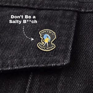 May include: A gold-colored enamel pin with the text "Don't Be a Salty B**ch" and an illustration of a person under an umbrella in the rain. The pin is attached to a black denim jacket.