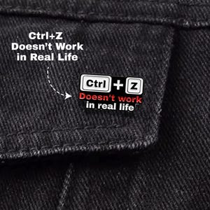 以下が含まれることがあります： 「Ctrl+Z Doesn't work in real life」のテキストが入った白黒のエナメルピン。ピンは、白と黒のデザインでCtrlキーとZキーを特徴とし、テキストは赤と白です。ピンは黒のデニムジャケットに取り付けられています。