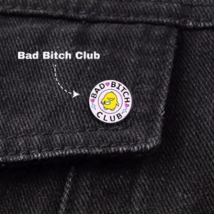 Puede incluir: Un pin redondo de esmalte rosa con las palabras "Bad Bitch Club" y un pollito de dibujos animados. El pin está sujeto a una chaqueta vaquera negra. El texto "Bad Bitch Club" también está escrito encima del pin.