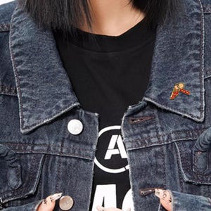 May include: A dark denim jacket over a black t-shirt. A small pin with a red, yellow, and white design is attached to the jacket. The pin has the text "New Day" on it.