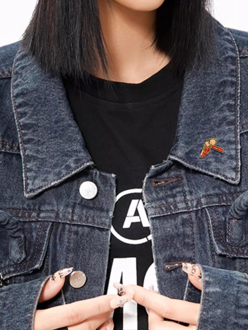 May include: A dark denim jacket over a black t-shirt. A small pin with a red, yellow, and white design is attached to the jacket. The pin has the text "New Day" on it.