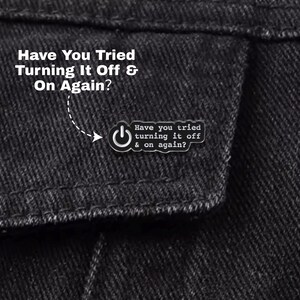 Könnte beinhalten: Nahaufnahme einer schwarzen Jeansjacke mit einer kleinen Anstecknadel. Die Anstecknadel zeigt ein Power-Button-Symbol und den Text "Have you tried turning it off & on again?" in Weiß. Die Jacke hat auch den gleichen Text in größerer Schrift.