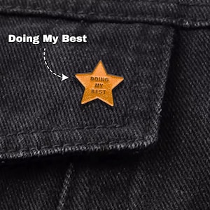 May include: A gold-colored star-shaped pin with the words "DOING MY BEST" in a dark font. The pin is attached to a black denim jacket. The text "Doing My Best" is above the pin.