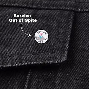 May include: A circular enamel pin featuring the words "Survive Out of Spite" and a floral design. The pin is white with a pink and blue flower illustration. The pin is attached to a black denim jacket.