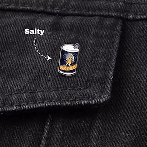 May include: A metal pin shaped like a can, with a cartoon character and the word "SALTY". The pin is fixed to a black denim jacket. The word "Salty" is written above the pin.