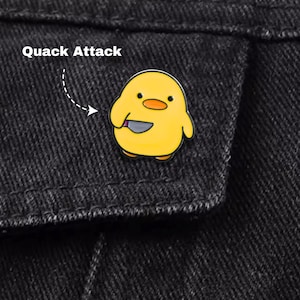 Può includere: Spilla in smalto giallo raffigurante un pulcino dei cartoni animati con un coltello. Il testo "Quack Attack" è visualizzato sopra la spilla, su uno sfondo di denim nero.