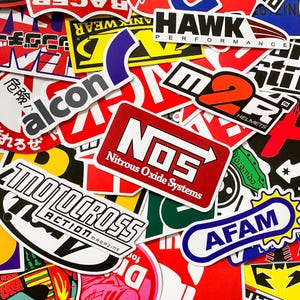 Könnte beinhalten: Eine Sammlung bunter Aufkleber mit verschiedenen Logos und Texten. Die Aufkleber zeigen Marken wie "NOS Nitrous Oxide Systems", "Motocross Action Magazine" und "AFAM". Die Farbpalette umfasst Rot, Weiß, Schwarz, Gelb und Blau.