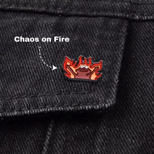 May include: A cartoon pin depicting a character in flames, with red and orange hues. The pin is attached to a black denim jacket. The text "Chaos on Fire" is above the pin.