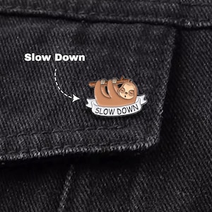 Pode incluir: Um alfinete de preguiça castanho com as palavras "SLOW DOWN" numa faixa branca. O alfinete está preso a uma jaqueta jeans preta. Acima do alfinete, as palavras "Slow Down" estão escritas em branco.