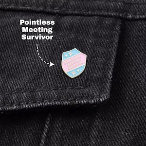 May include: A colorful enamel pin with the text "Pointless Meeting Survivor" on a black denim jacket. The pin is shield-shaped with a light blue and pink color scheme and star accents.