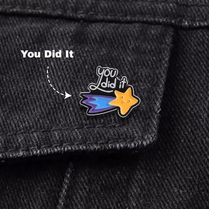 May include: A black denim jacket with a colorful enamel pin. The pin features a yellow star with a smiling face, a blue and purple comet tail, and the words "You Did It". The text "You Did It" is also printed above the pin.