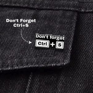 以下が含まれることがあります： 「Don't Forget Ctrl+S」の文字が入った白黒のエナメルピン。ピンには「Ctrl + S」の文字が入った白い長方形と黒い背景があります。ピンは黒いデニムジャケットに取り付けられています。